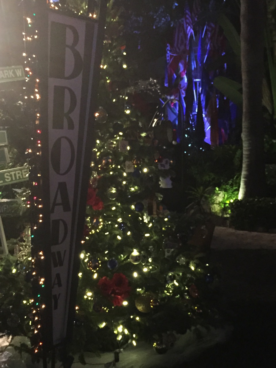 A lit holiday tree beside a vertical Broadway sign in a garden.