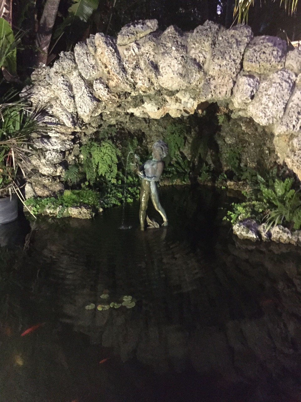 A decorative mermaid figure beneath a rock grotto above a dark pond.