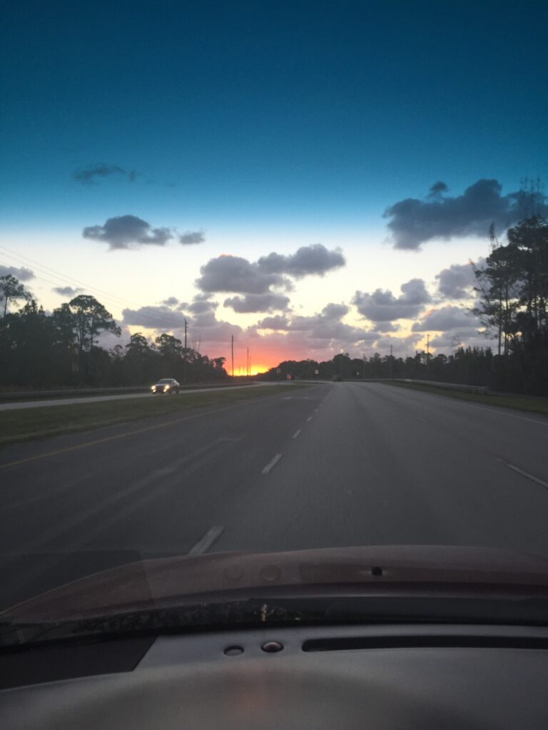 View through a windshield of a road, scattered clouds, and an orange morning horizon.