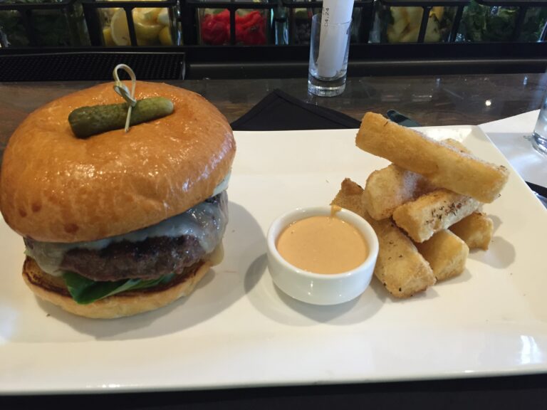 A burger with a pickle, toast sticks, and dipping sauce on a white plate.