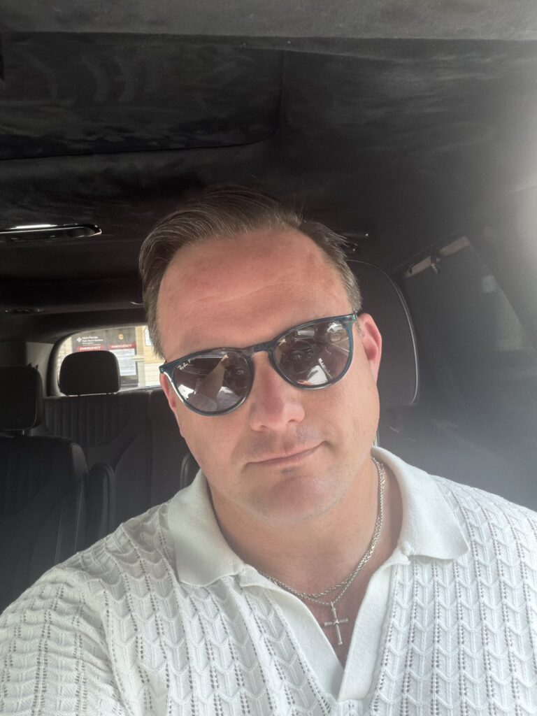 Portrait inside a car, wearing sunglasses and a white textured polo shirt.