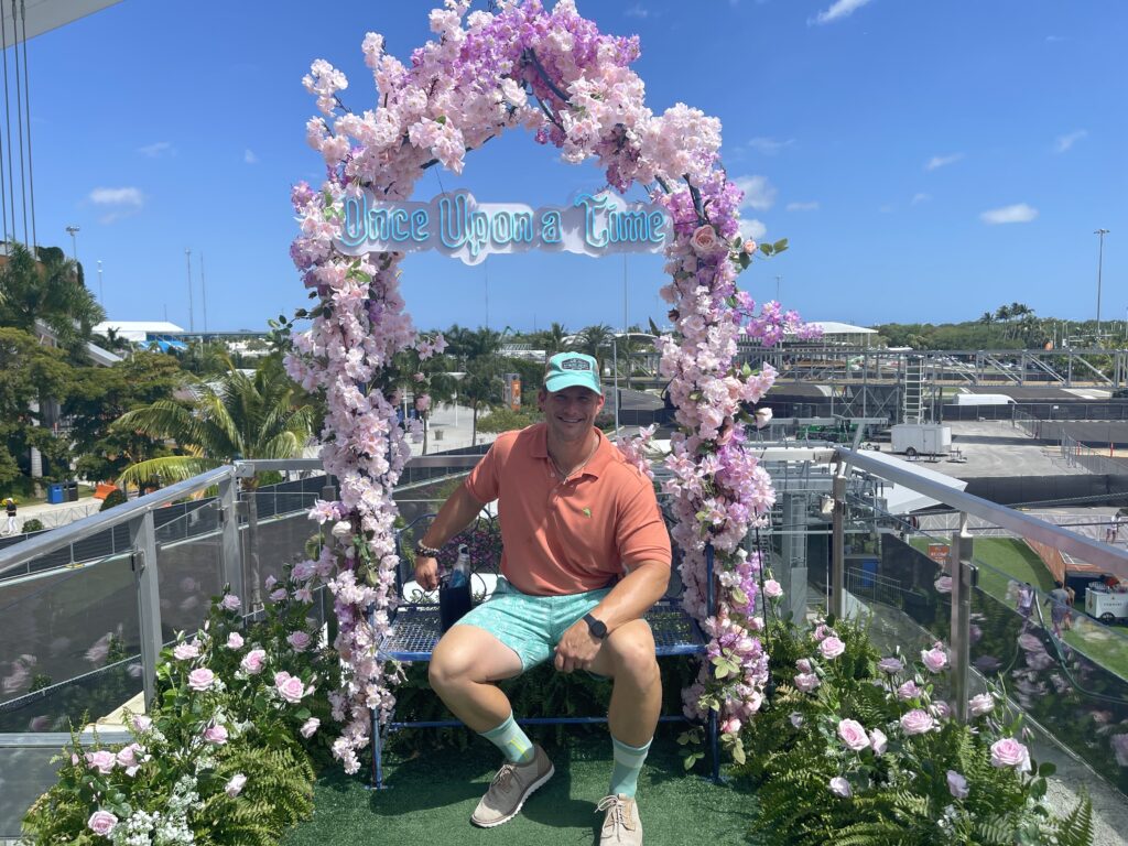 Man seated beneath a floral arch at an outdoor event.