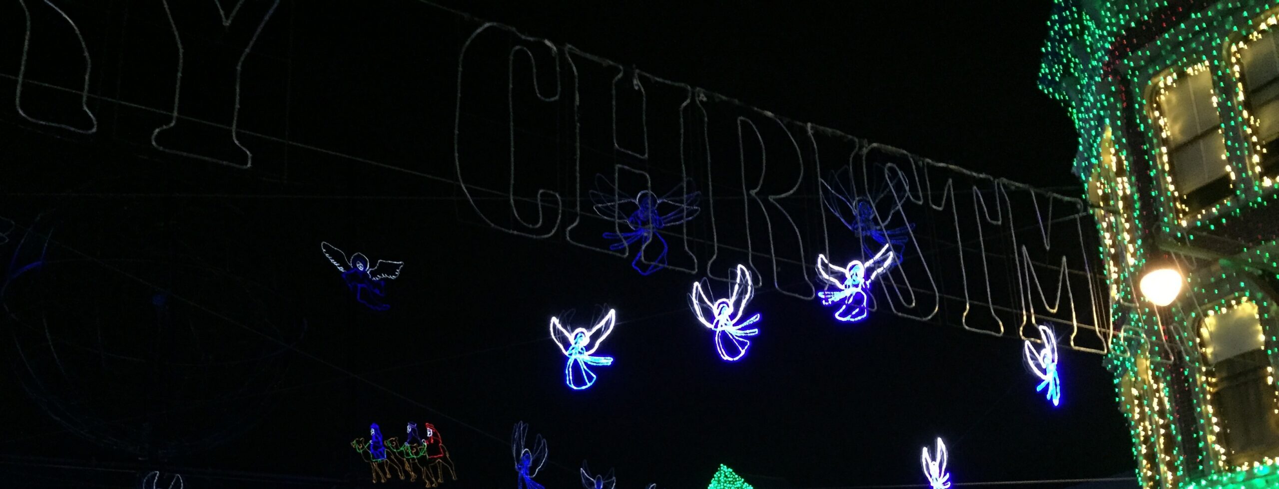 Illuminated angels suspended above a holiday-light street beside a building covered in green and white lights.