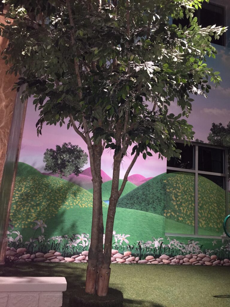 An illuminated tree beside a painted landscape wall at night.