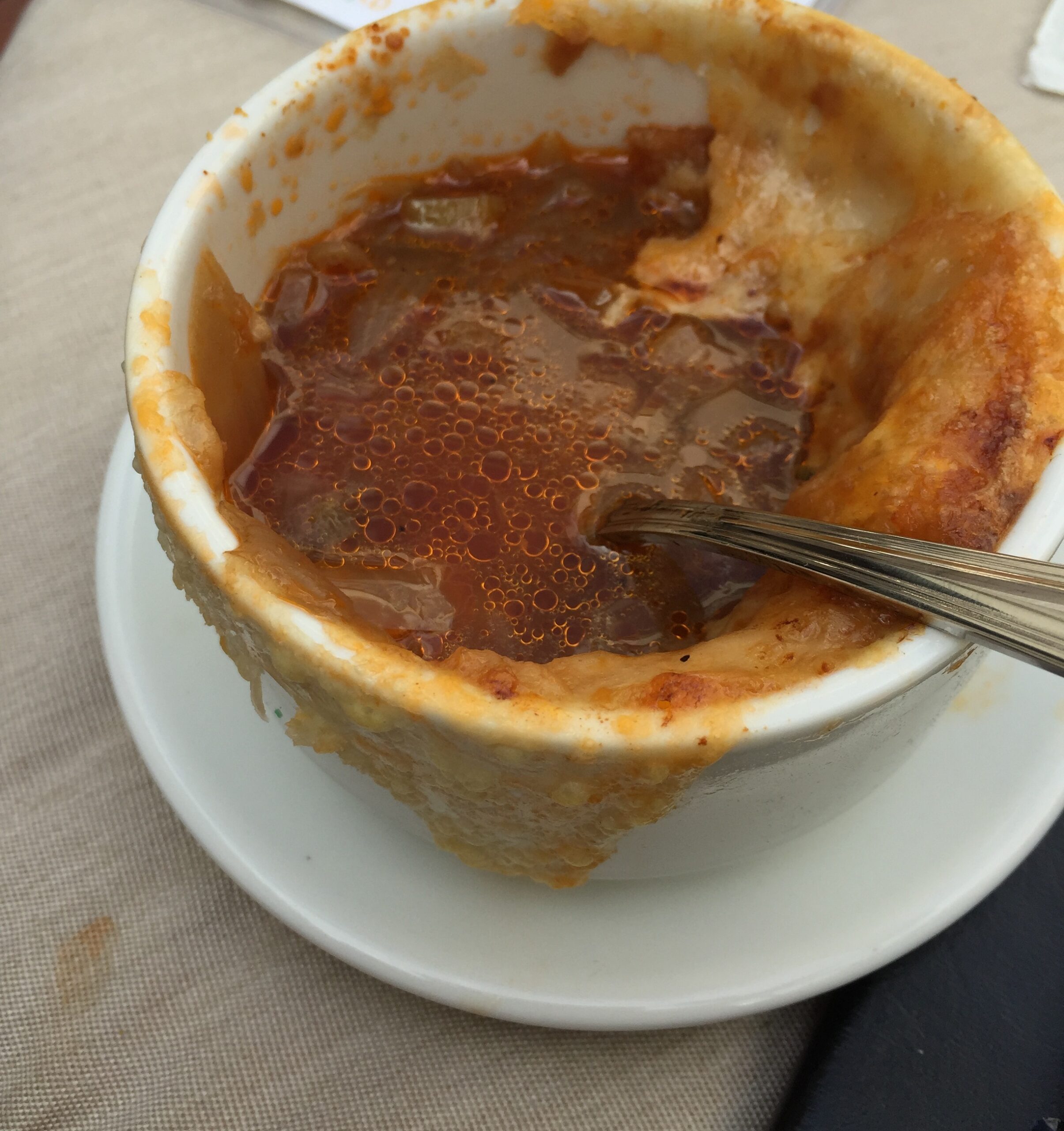 A bowl of brown soup with a browned cheese rim and a spoon.