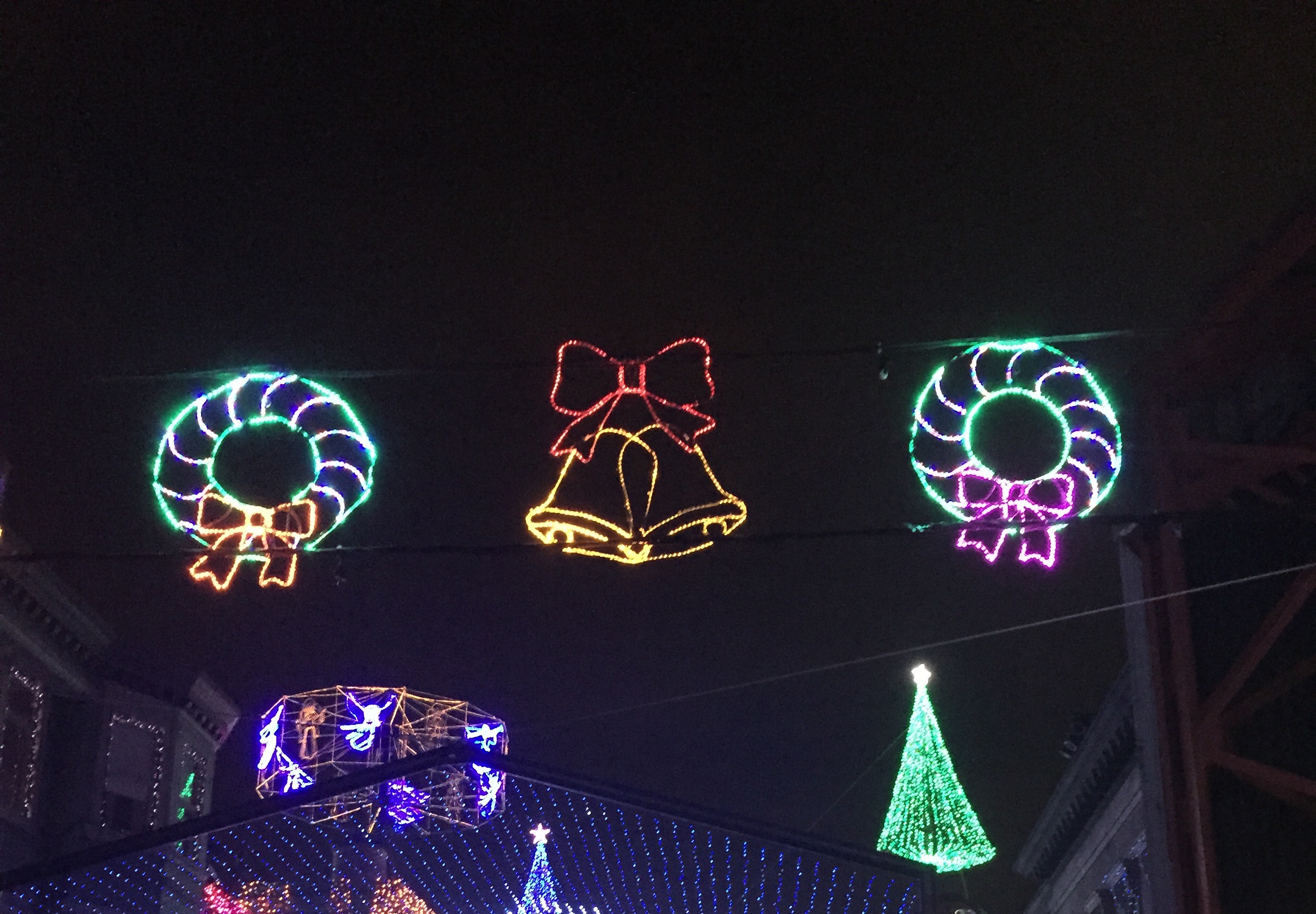 Upper view of a Disney street covered in multicolored holiday lights beneath illuminated wreaths and bells.