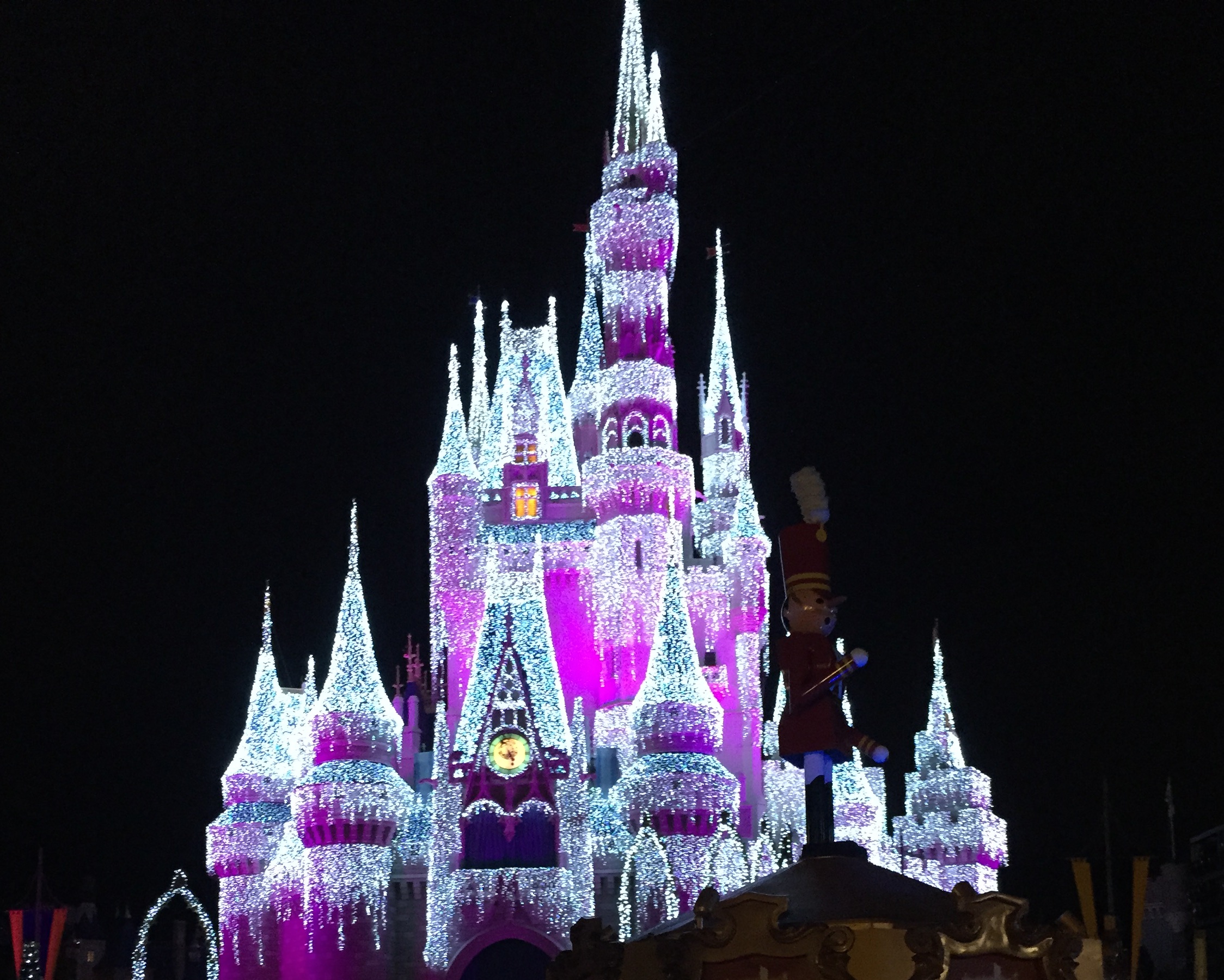 Cinderella Castle glowing in white and magenta lights beside a toy-soldier parade display.